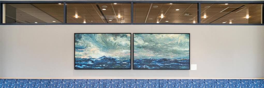 A diptych of moody blue and white waves and a teal and blue and pink sky. Looks like a storm. Paintings are hgning on a gray wall with windows and a long blue bench underneath.